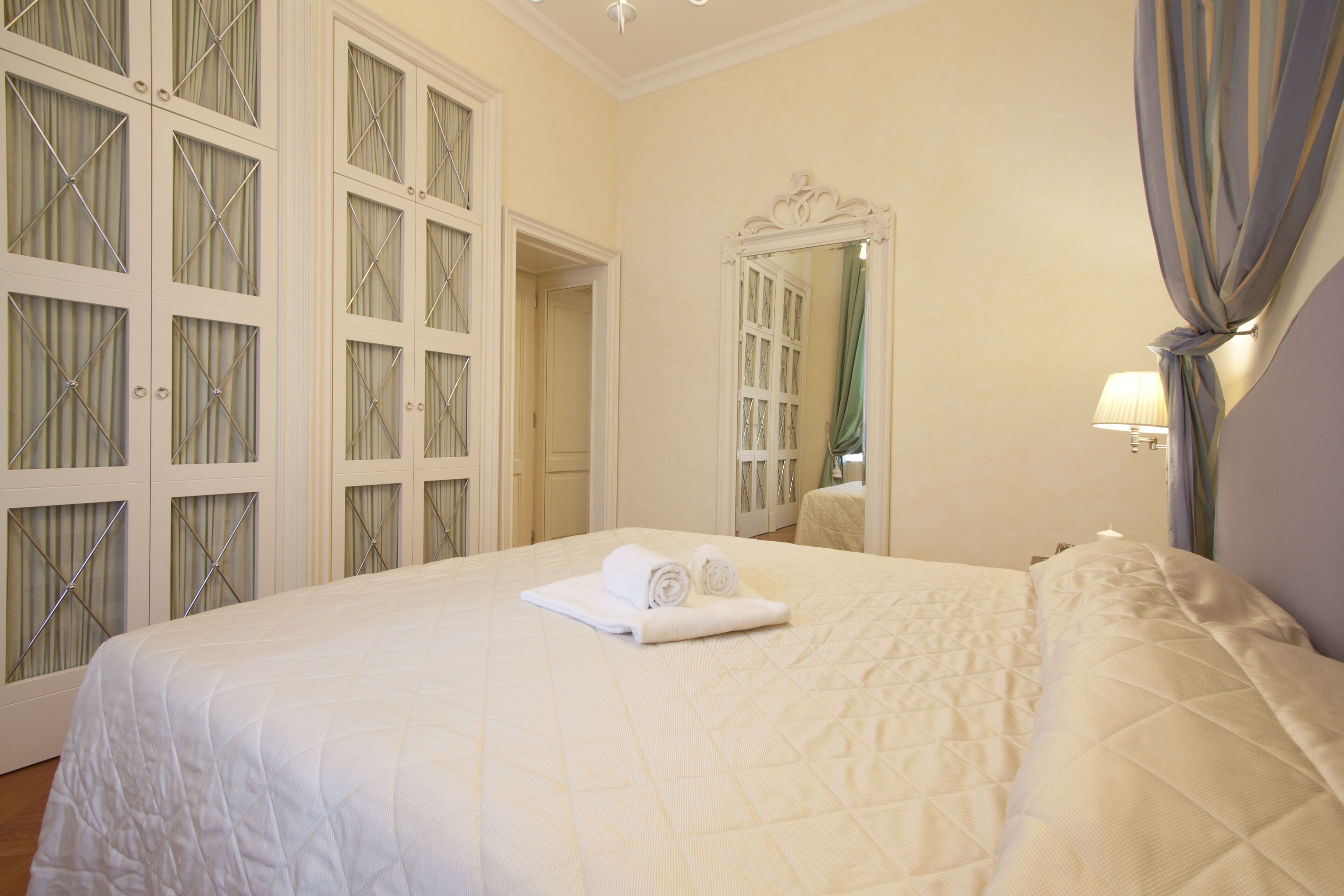 A hotel room with a large bed, a wardrobe with mirrored doors, a bedside table with a lamp, and a mirror on the wall.