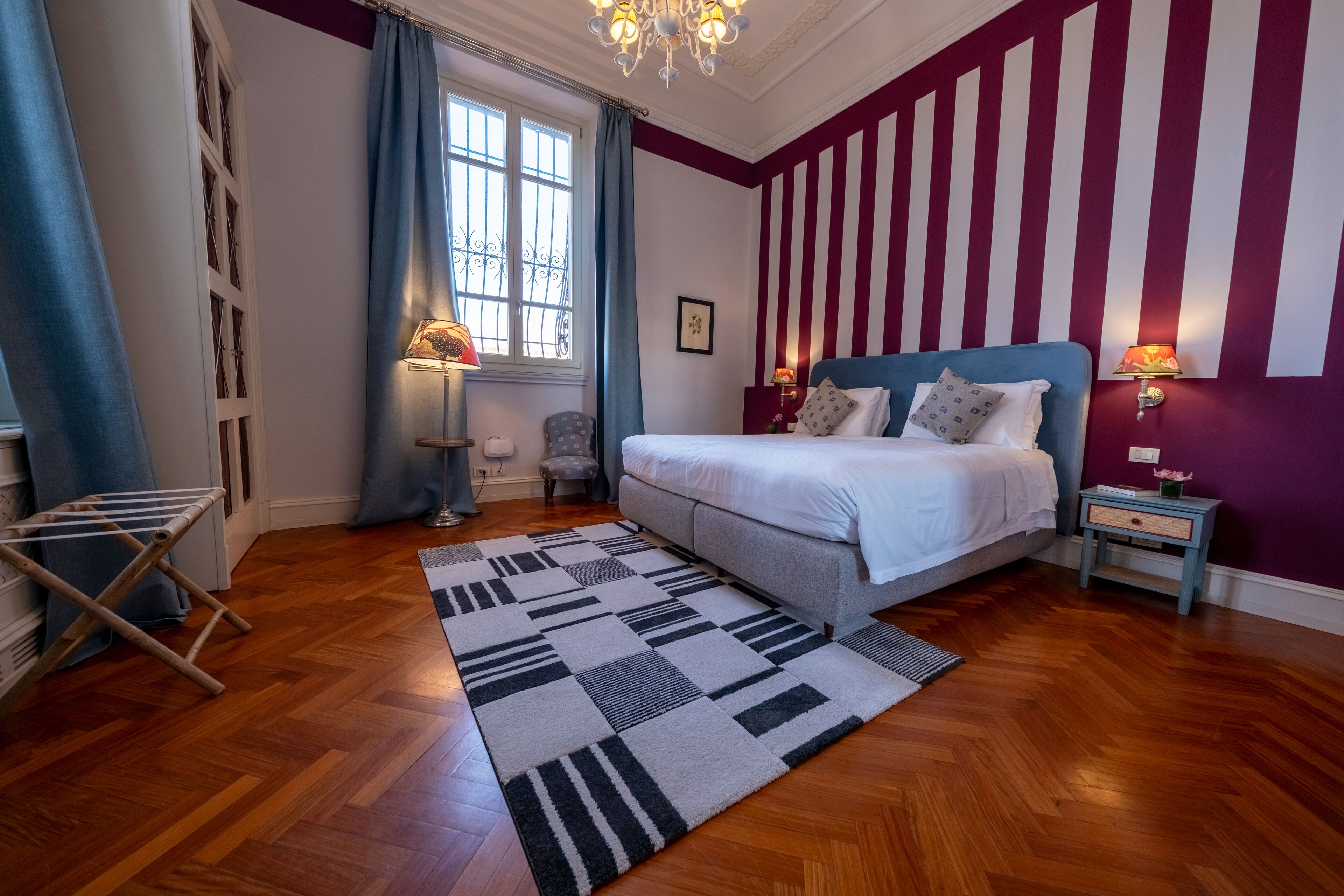 A bedroom with a bed, a window with curtains, a chandelier, and a striped wall.