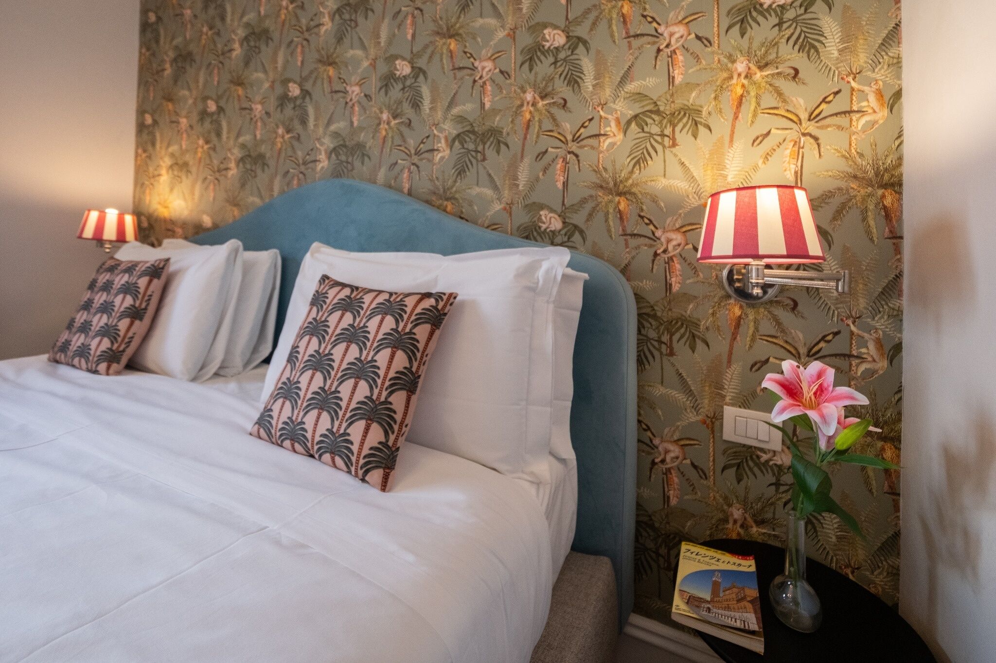 Une chambre d’hôtel avec un lit, un papier peint aux motifs tropicaux, une table de chevet avec une lampe et un vase avec une fleur.