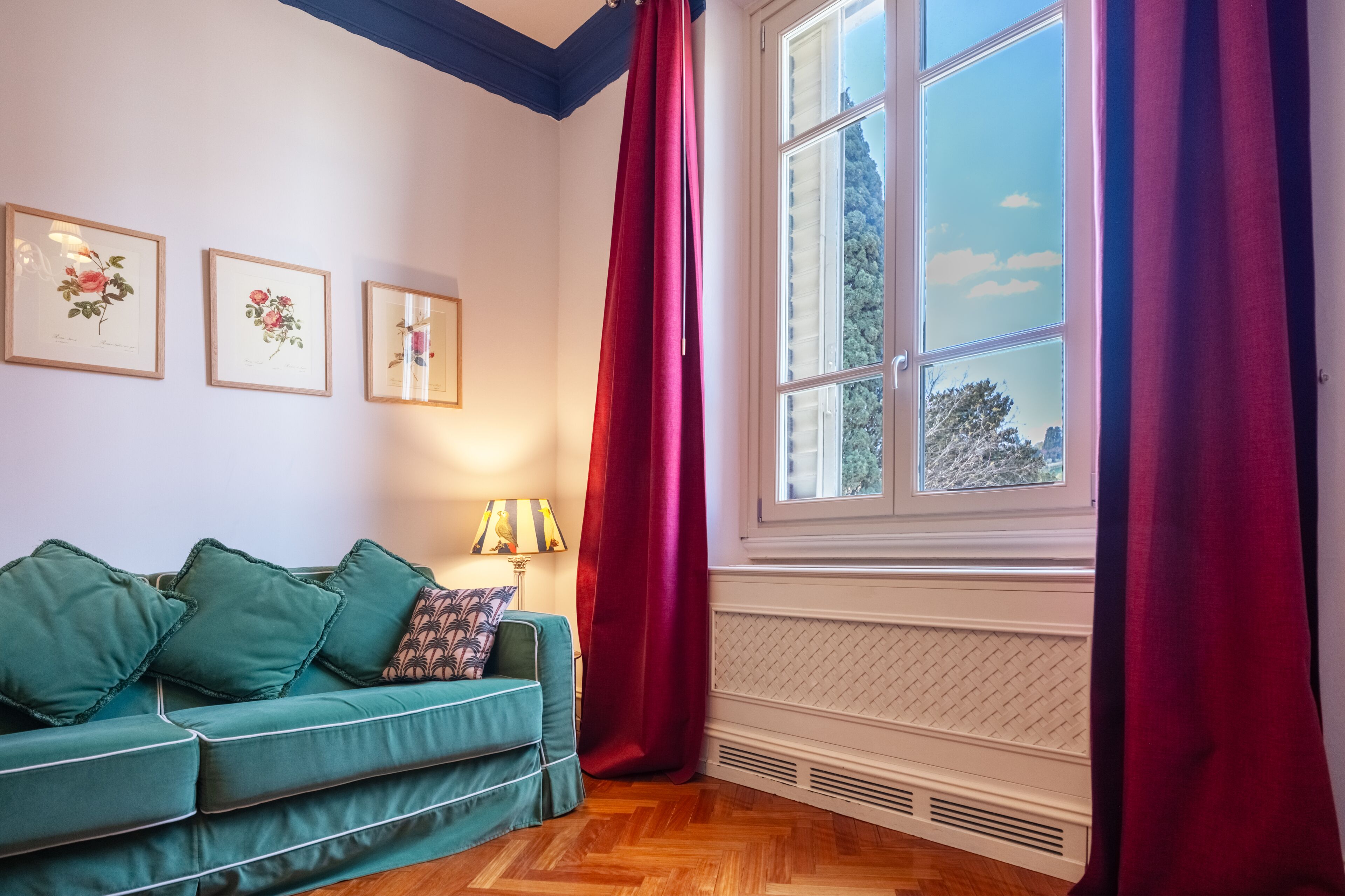 A room with a teal sofa, red curtains, and framed botanical prints on the wall.
