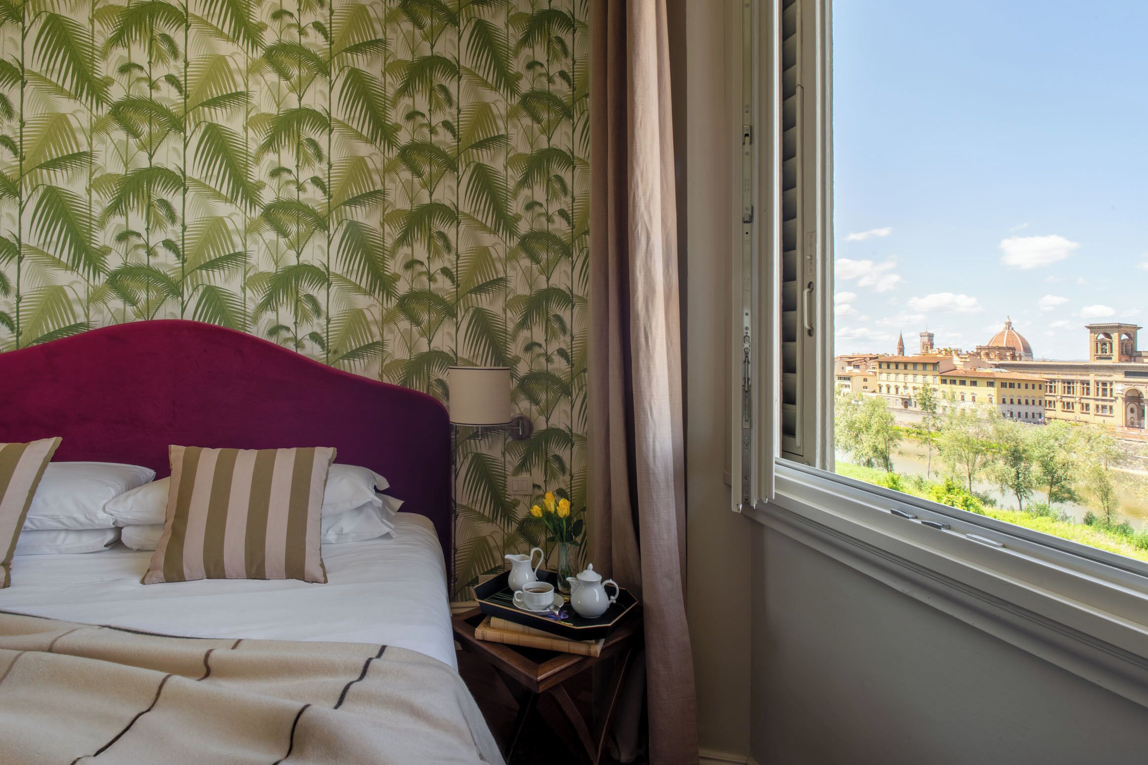 Ein Hotelzimmer mit einem Bett, einem Tablett mit Teekanne und Tassen, einer Vase mit Blumen und Blick auf eine Stadtlandschaft durch ein Fenster.