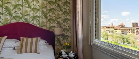 A hotel room with a bed, a tray with a teapot and cups, a vase with flowers, and a view of a cityscape through a window.