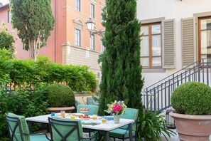 Un área de comedor al aire libre con una mesa para dos, rodeada de sillas, una planta en maceta y vistas a un edificio con una escalera.