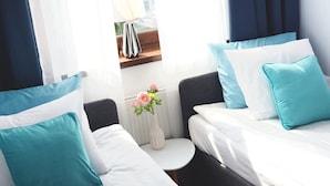 Habitación de hotel con dos camas, cada una con almohadas blancas y turquesa, una mesita blanca con un florero y una ventana con cortinas.