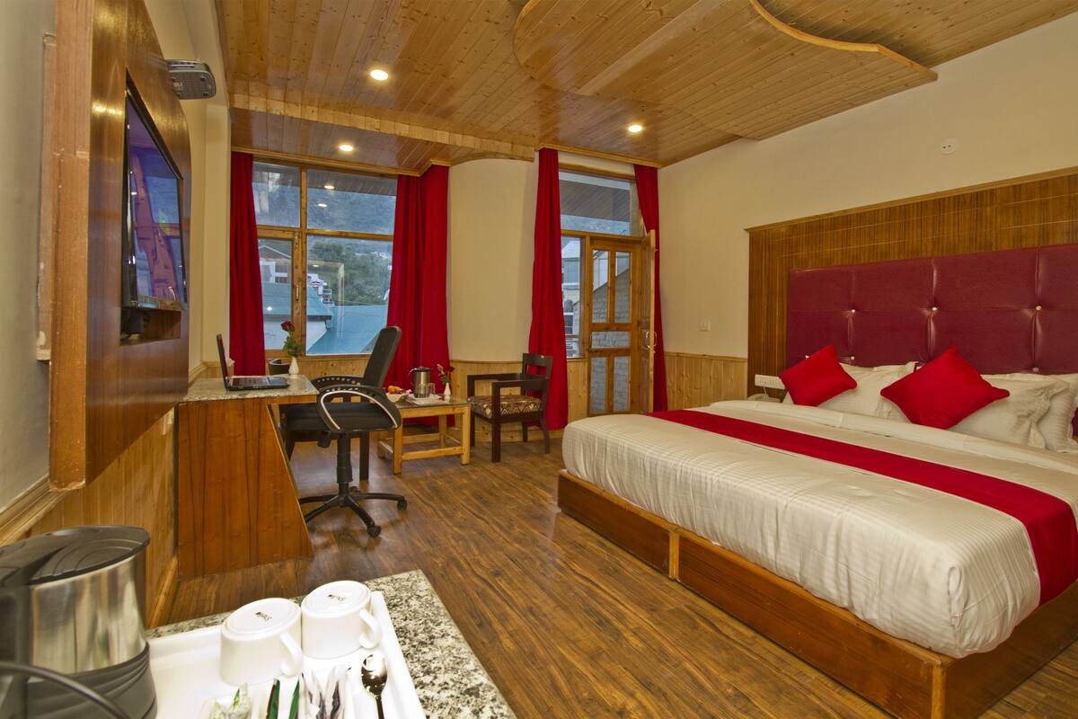 Habitación de hotel con techo de madera, una cama grande con almohadas rojas, un escritorio con silla y una ventana con cortinas rojas.