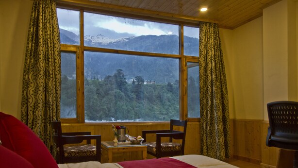 A room with a large window offering a mountain view, a bed with red pillows, two wooden chairs, and a small table with a vase and a book. - Sarthak Regency (Manali)