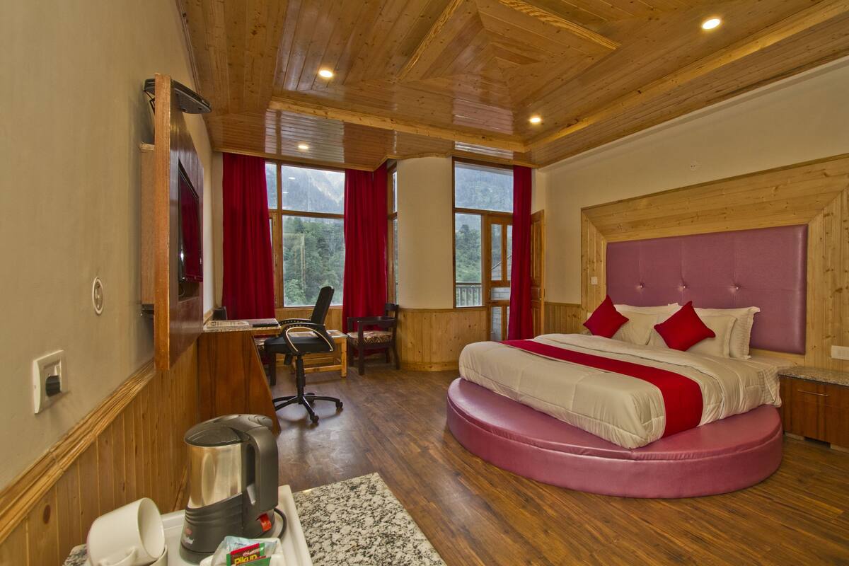 Un dormitorio con techo de madera, una cama grande con almohadas rojas, un escritorio con una silla y vistas a las montañas a través de las ventanas.