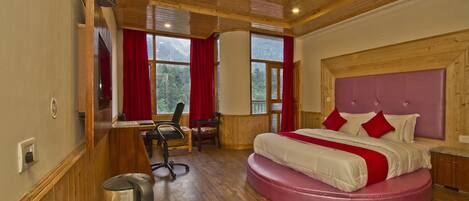 A bedroom with a wooden ceiling, a large bed with red pillows, a desk with a chair, and a view of mountains through the windows.