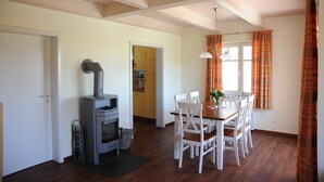 Een knus vertrek met een houten plafond, een eettafel met stoelen, een open haard en een keuken in de achtergrond.