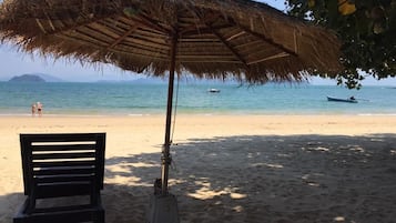A beach scene with a thatched umbrella, a wooden bench, and a calm sea with boats in the distance.