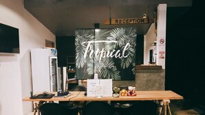 Un comptoir d’accueil surmonté d’une enseigne indiquant « Réception », une autre enseigne avec des feuilles de palmier indiquant « Bienvenue », un menu posé sur la table et trois chaises noires devant.