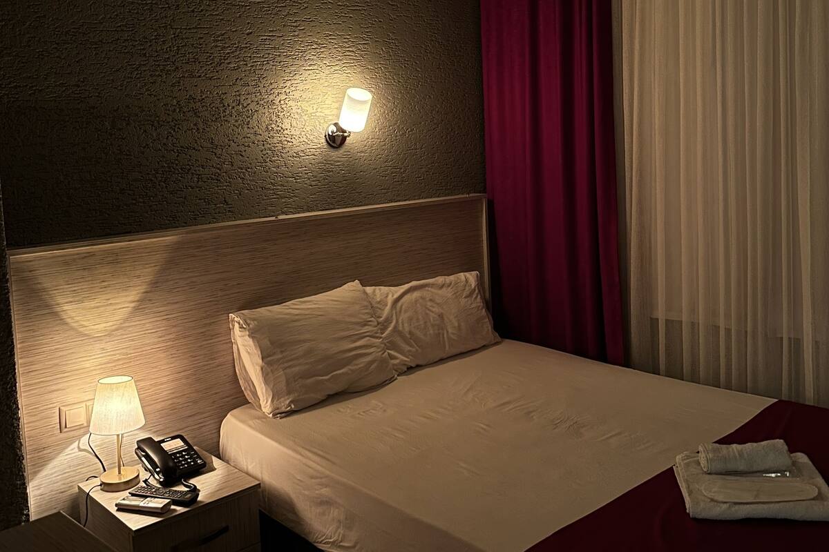 A hotel room with a bed, a bedside table with a lamp and phone, a wall-mounted light, and a window with curtains.