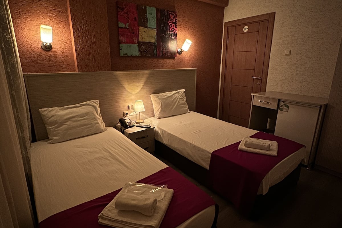 A hotel room with two single beds, a nightstand with a lamp, a wall-mounted artwork, and a door with a sign.