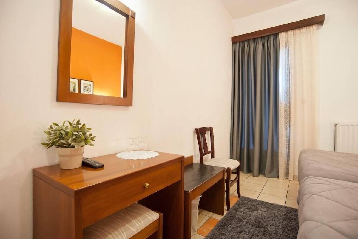 Un dormitorio con cama, mesita de noche con una planta, una silla, escritorio con espejo y ventana con cortinas.