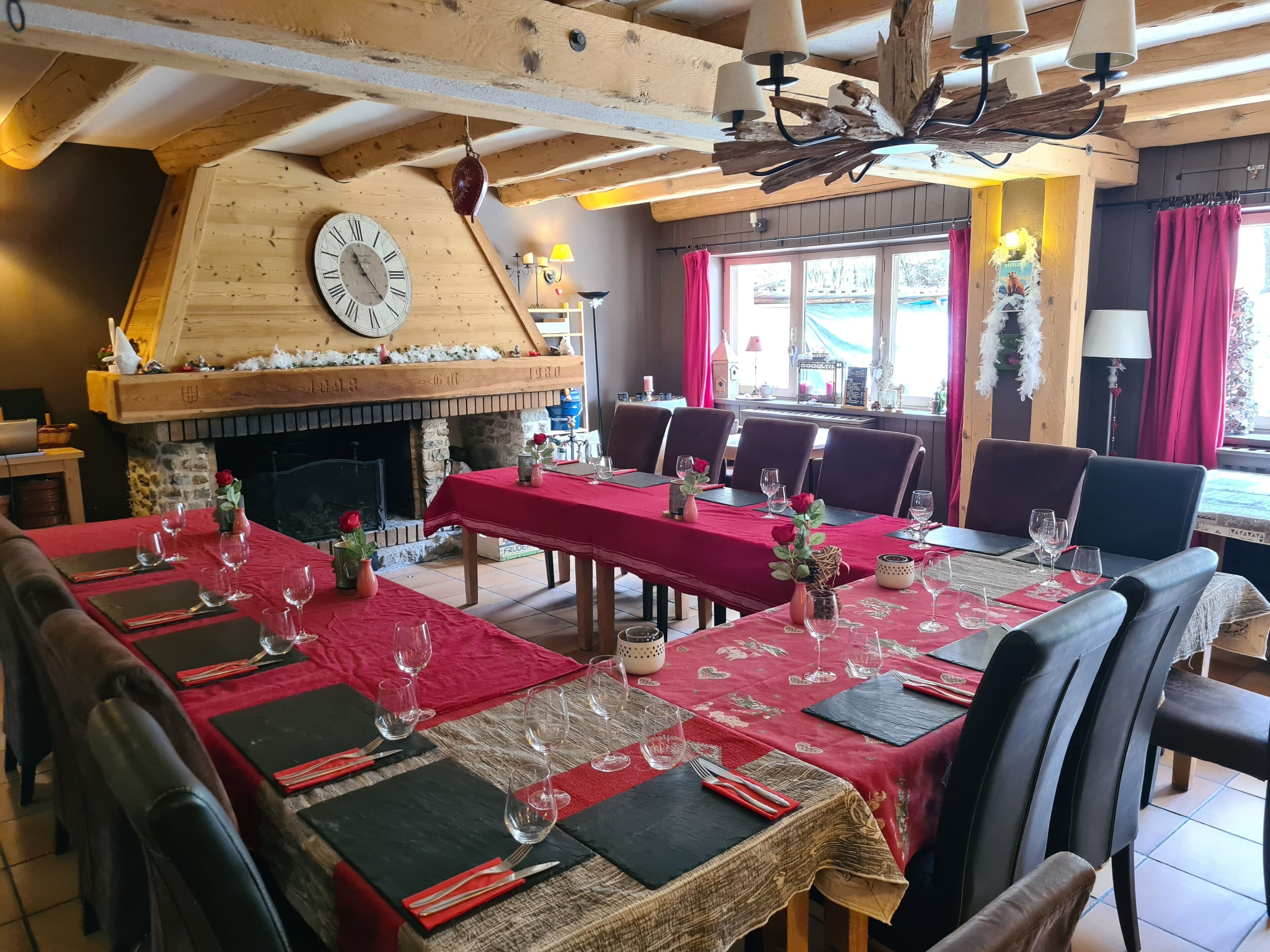 A dining area with a wooden ceiling, a fireplace, and a clock. Tables are set with red tablecloths and wine glasses.