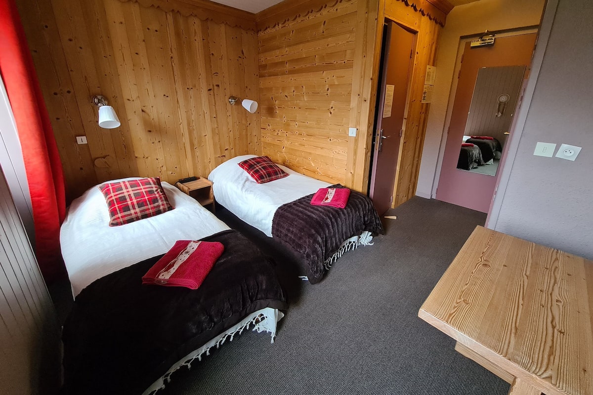 A room with two beds, each with a red and black checkered pillow, a small wooden nightstand, and a table.