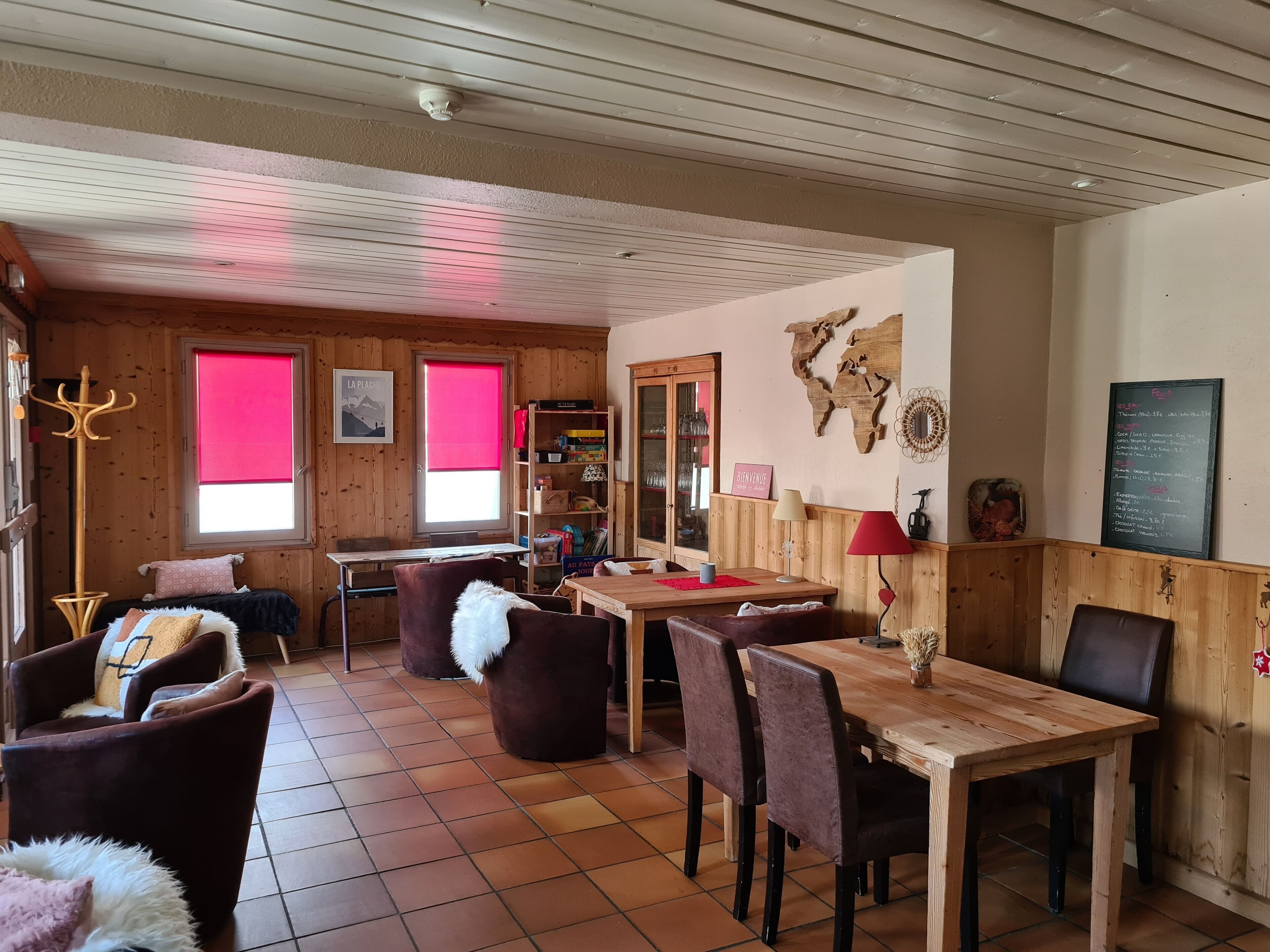 A cozy room with wooden walls, a dining table, chairs, a sofa, and a window with a red blind.