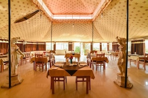Uma sala de jantar com mesas preparadas para o serviço, uma estátua de mulher à esquerda e uma estátua de homem à direita.