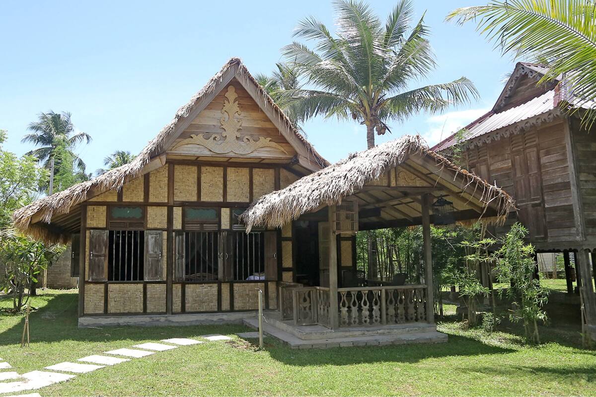 A traditional wooden house with a thatched roof, surrounded by a well-maintained garden and palm trees.