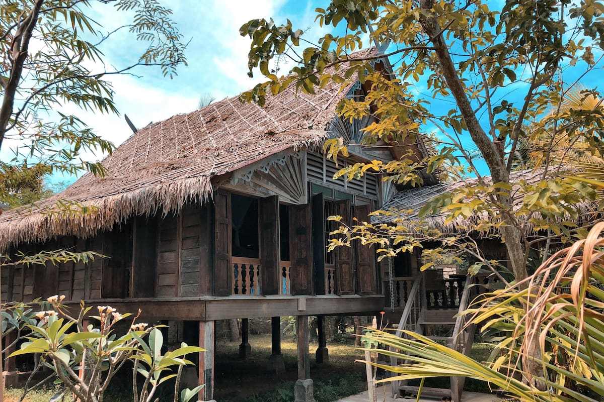 A traditional wooden house with a thatched roof, elevated on stilts, surrounded by tropical vegetation.