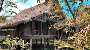 A traditional wooden house with a thatched roof, elevated on stilts, surrounded by tropical vegetation.