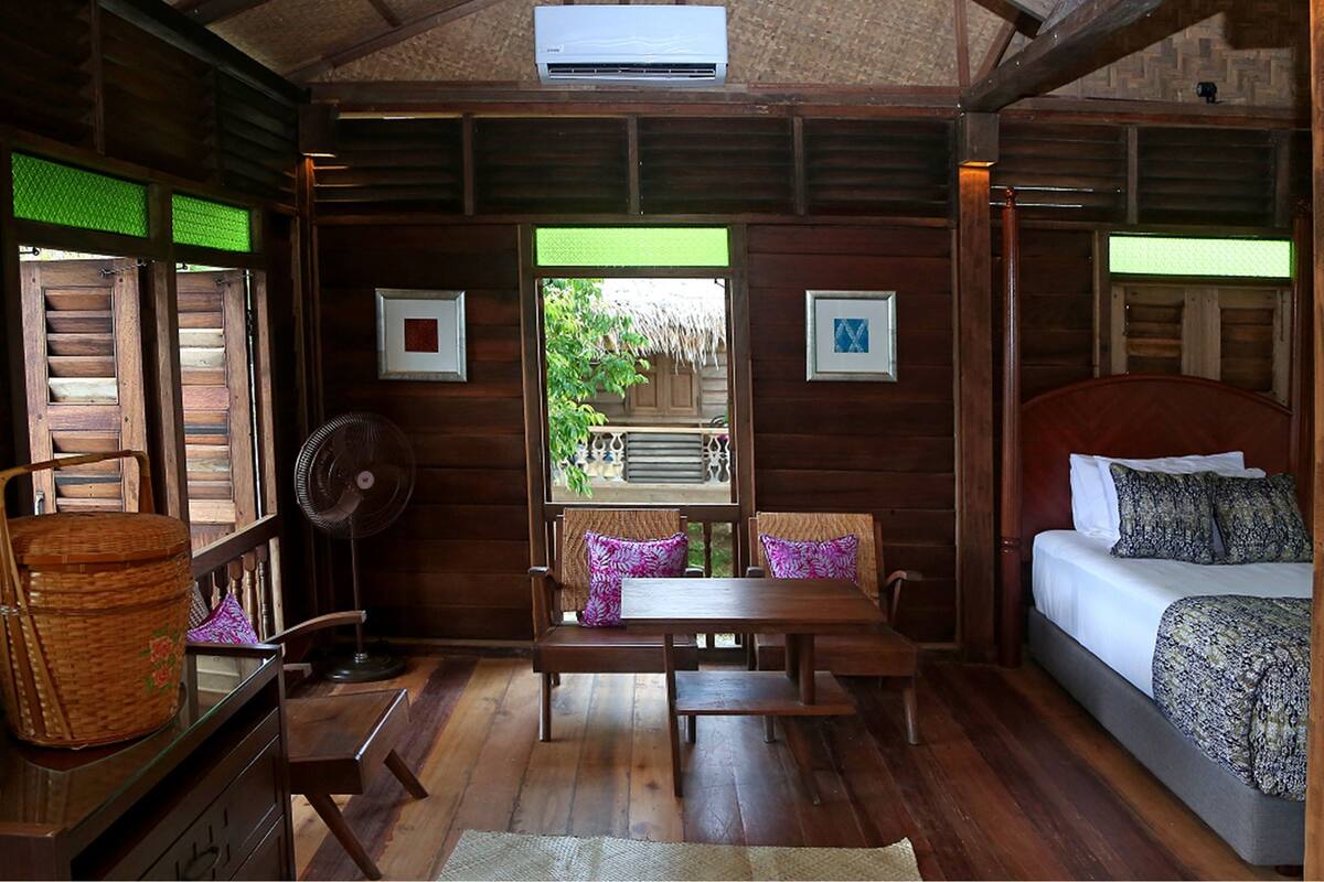 A wooden cabin room with a bed, a table, chairs, a fan, and a window with a view of greenery.