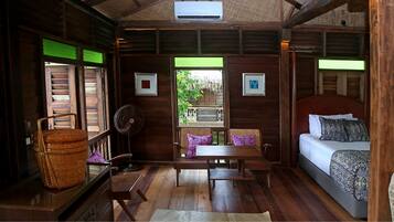 A wooden cabin room with a bed, a table, chairs, a fan, and a window with a view of greenery.