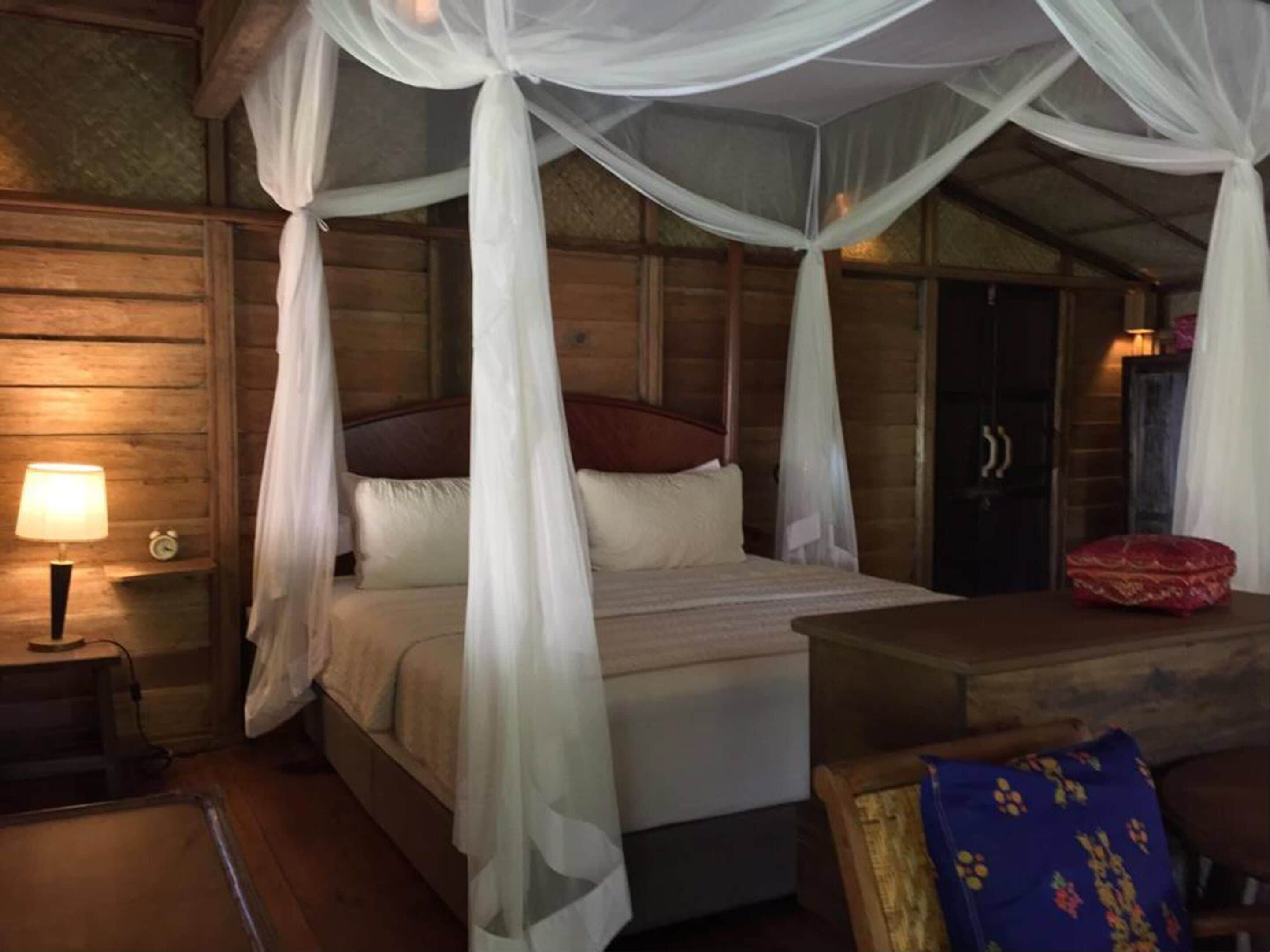 A wooden room with a bed under a canopy, a nightstand with a lamp, and a small table with a red cushion.
