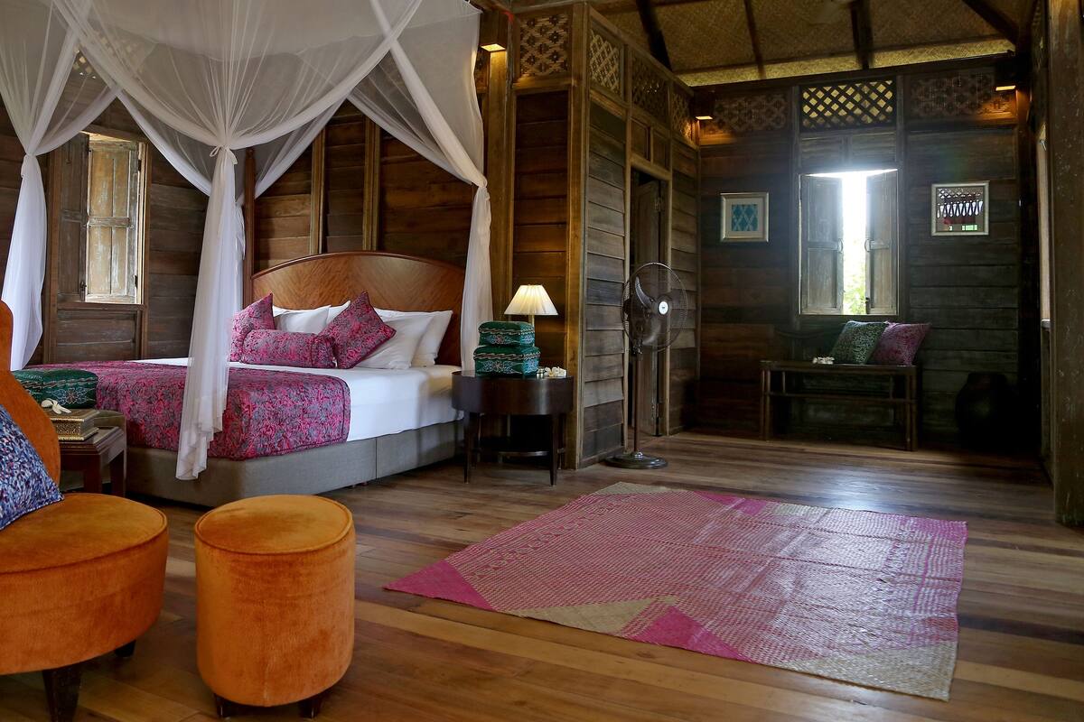 A wooden room with a bed, a chair, ottomans, a fan, and a rug.