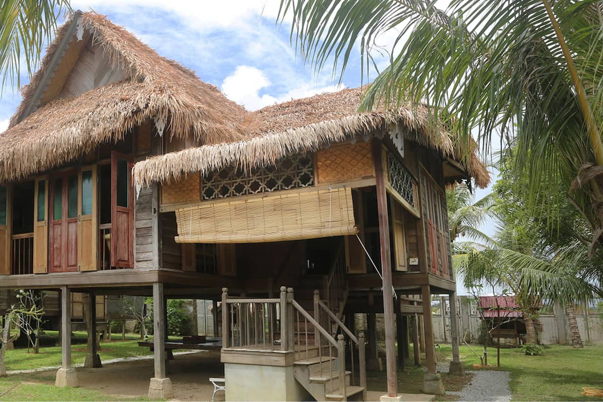 A traditional wooden house with a thatched roof, a balcony, and a staircase leading to the upper level.