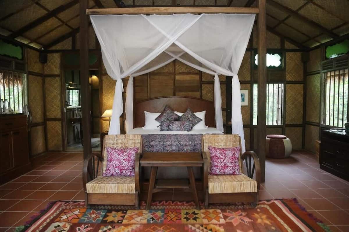 A room with a four-poster bed, two woven chairs, a small table, and a patterned rug.
