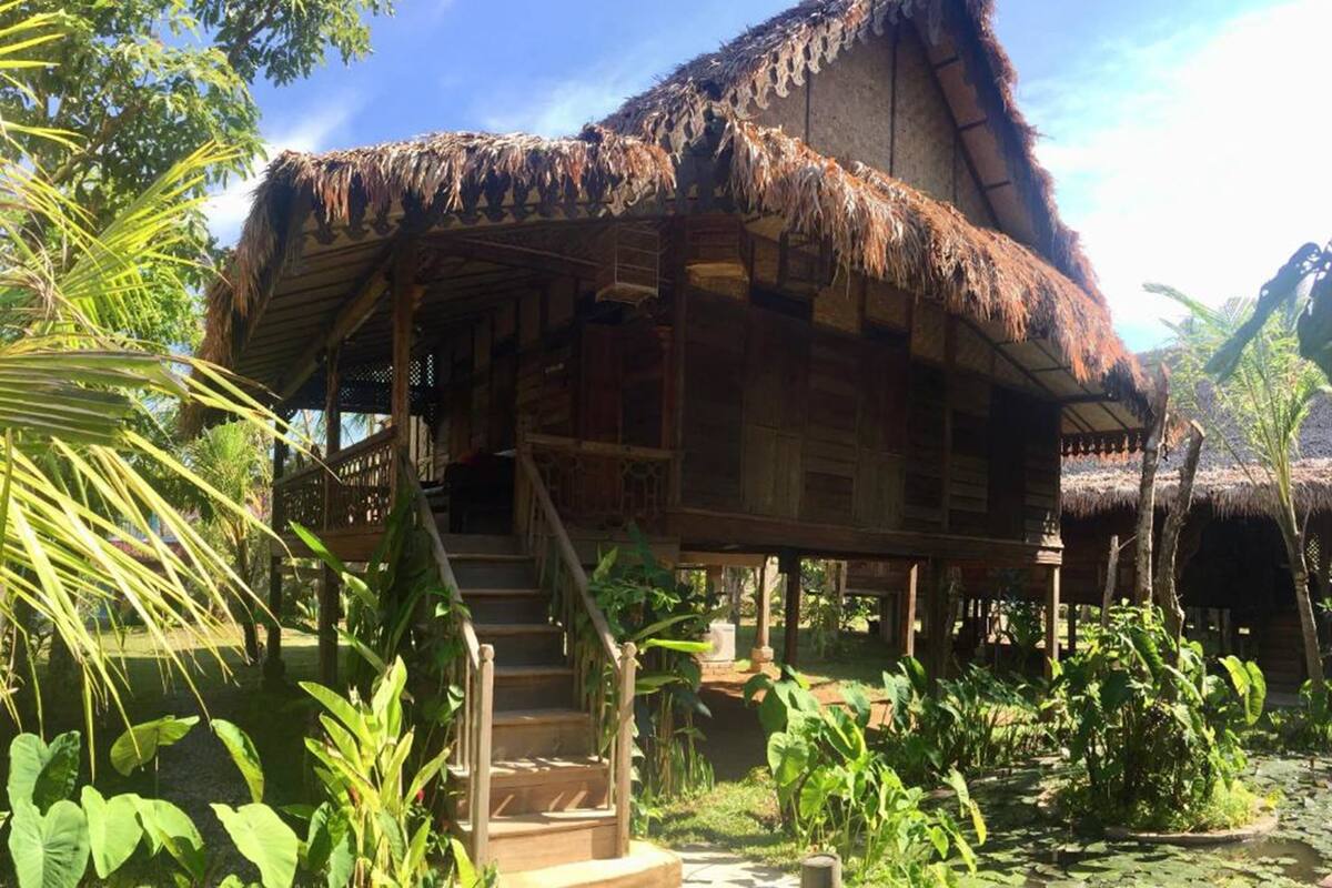 A rustic wooden house with a thatched roof, surrounded by lush greenery and a clear sky.