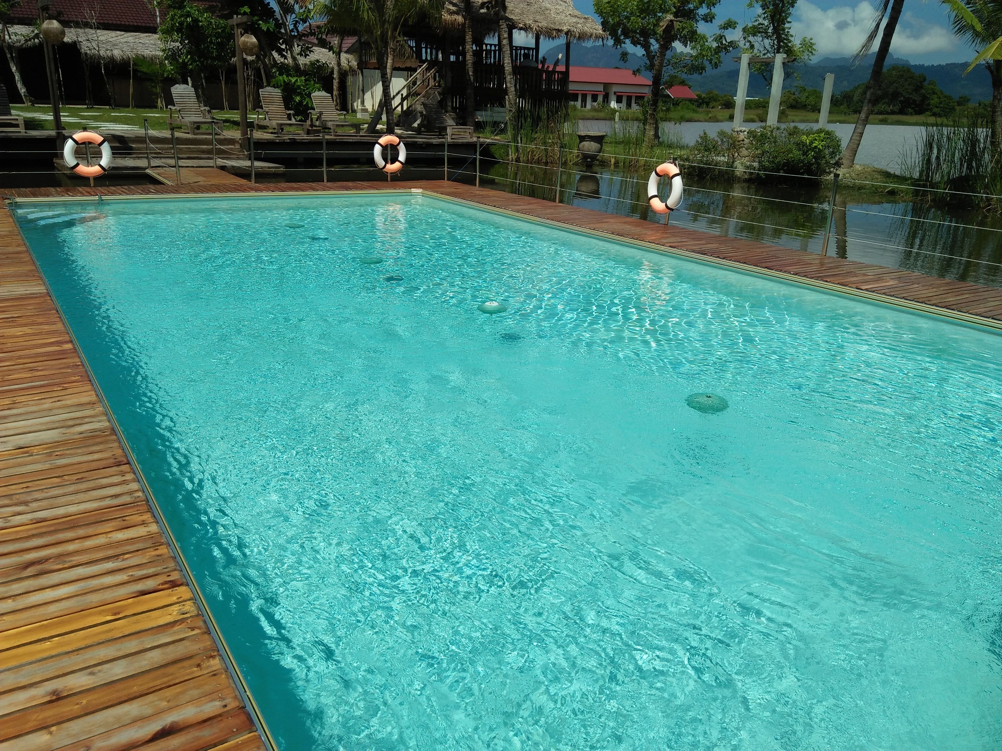 Una piscina rectangular con agua cristalina, rodeada por una terraza de madera. Se observan chalecos salvavidas en el borde de la piscina.