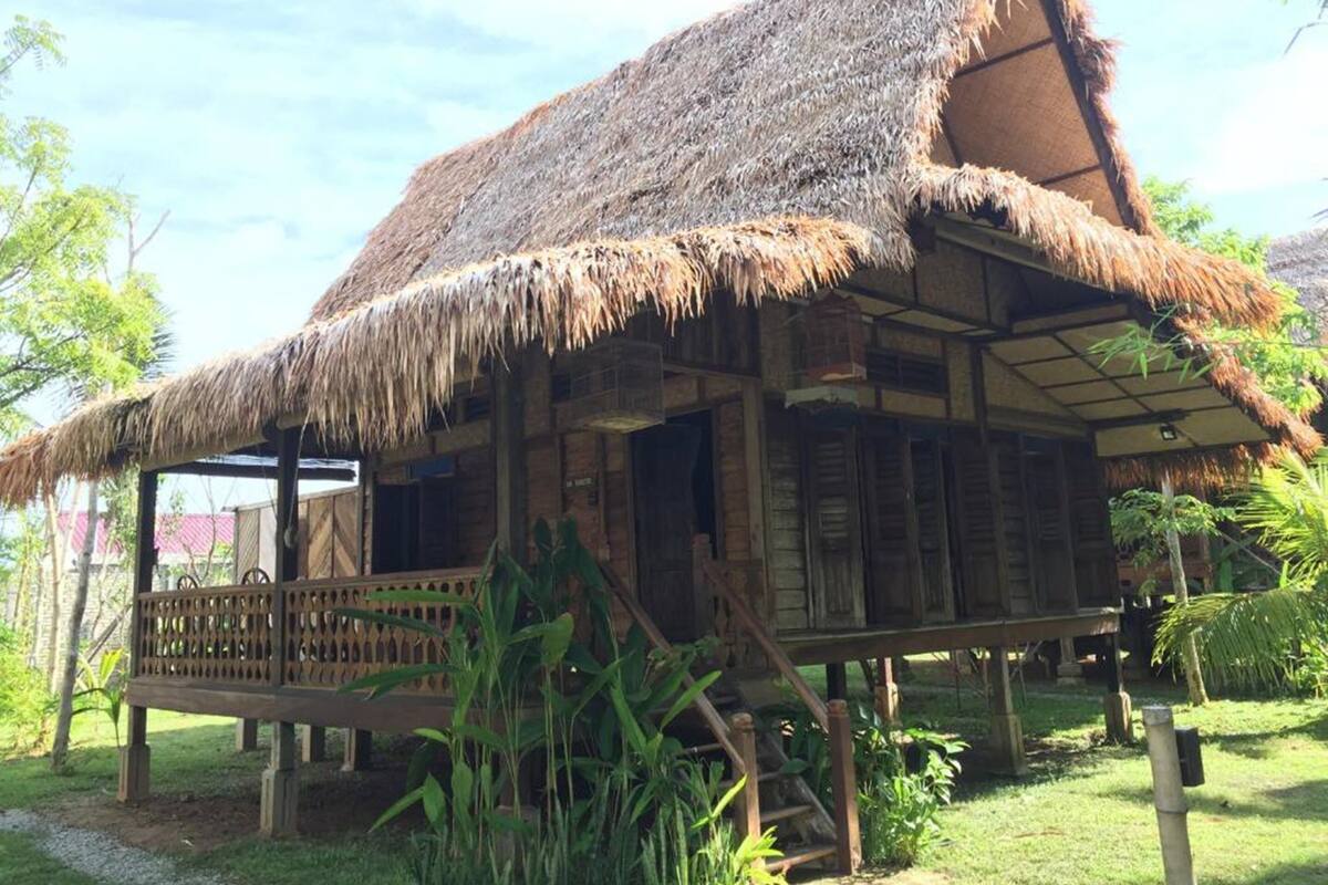 A traditional thatched-roof house with a wooden deck and surrounding greenery.