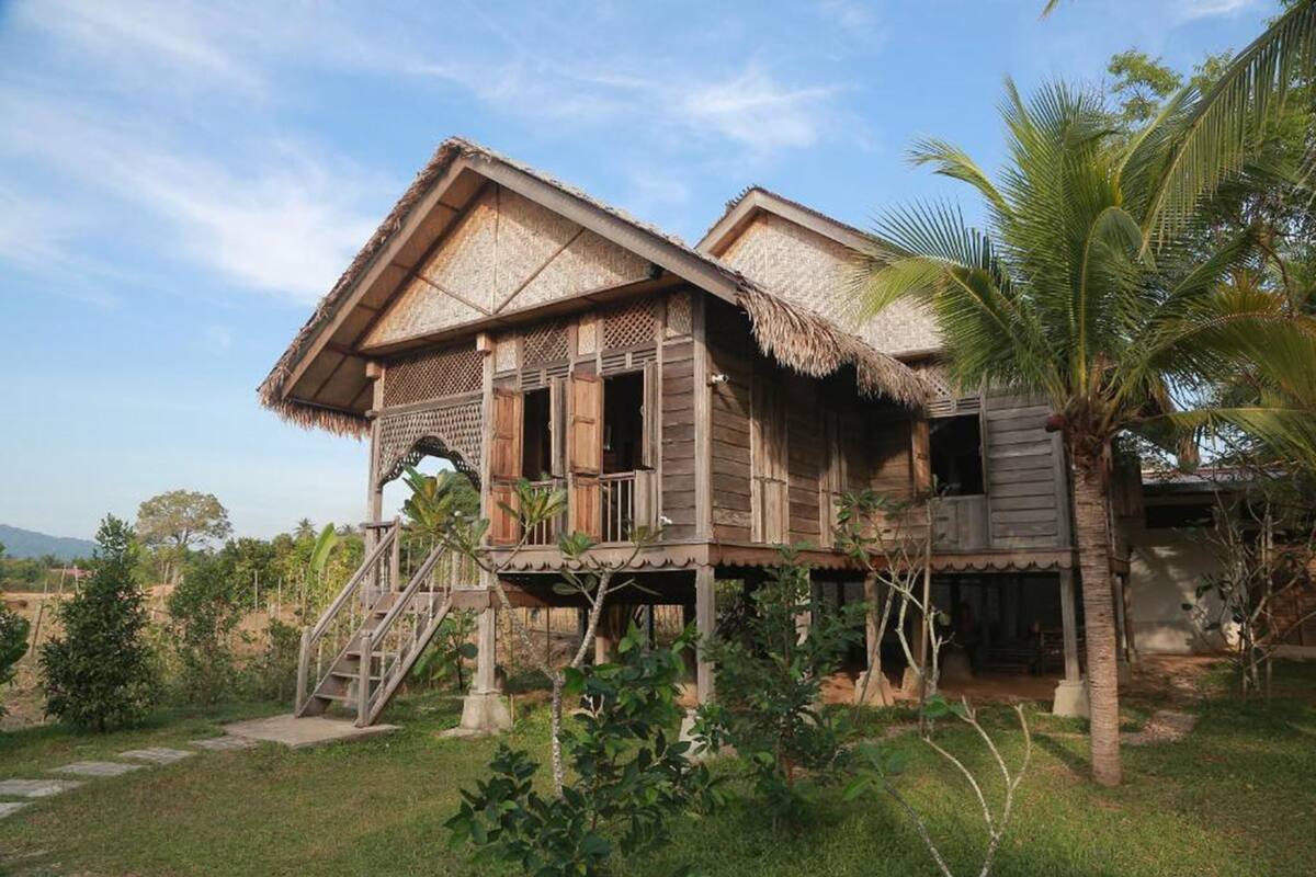 A wooden house with a thatched roof, a balcony, and a staircase leading to the entrance.