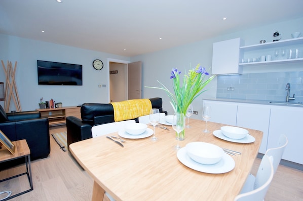 A modern living room with a wooden dining table set for four, a black leather sofa, and a yellow throw.