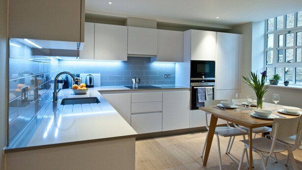 A modern kitchen with white cabinetry, a central island with a sink, and a dining area with a table set for two. - The London Agent Spitalfields (London)