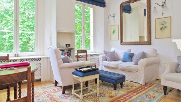 A cozy living room with two white sofas, a blue ottoman, and a wooden table with a tray. Large windows with blue curtains allow natural light to fill the room. - The London Agent Hyde Park King Bed (London)