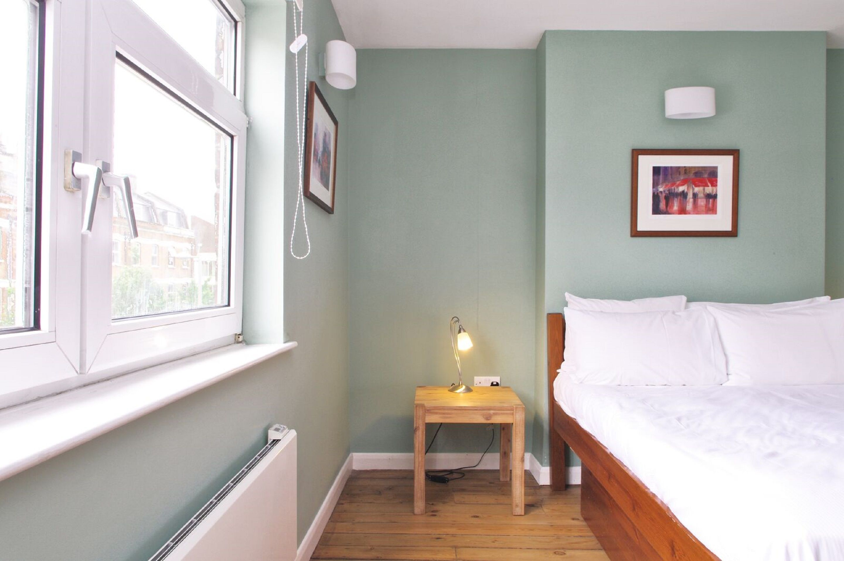 A room with a bed, a small table with a lamp, a window, and a picture on the wall.