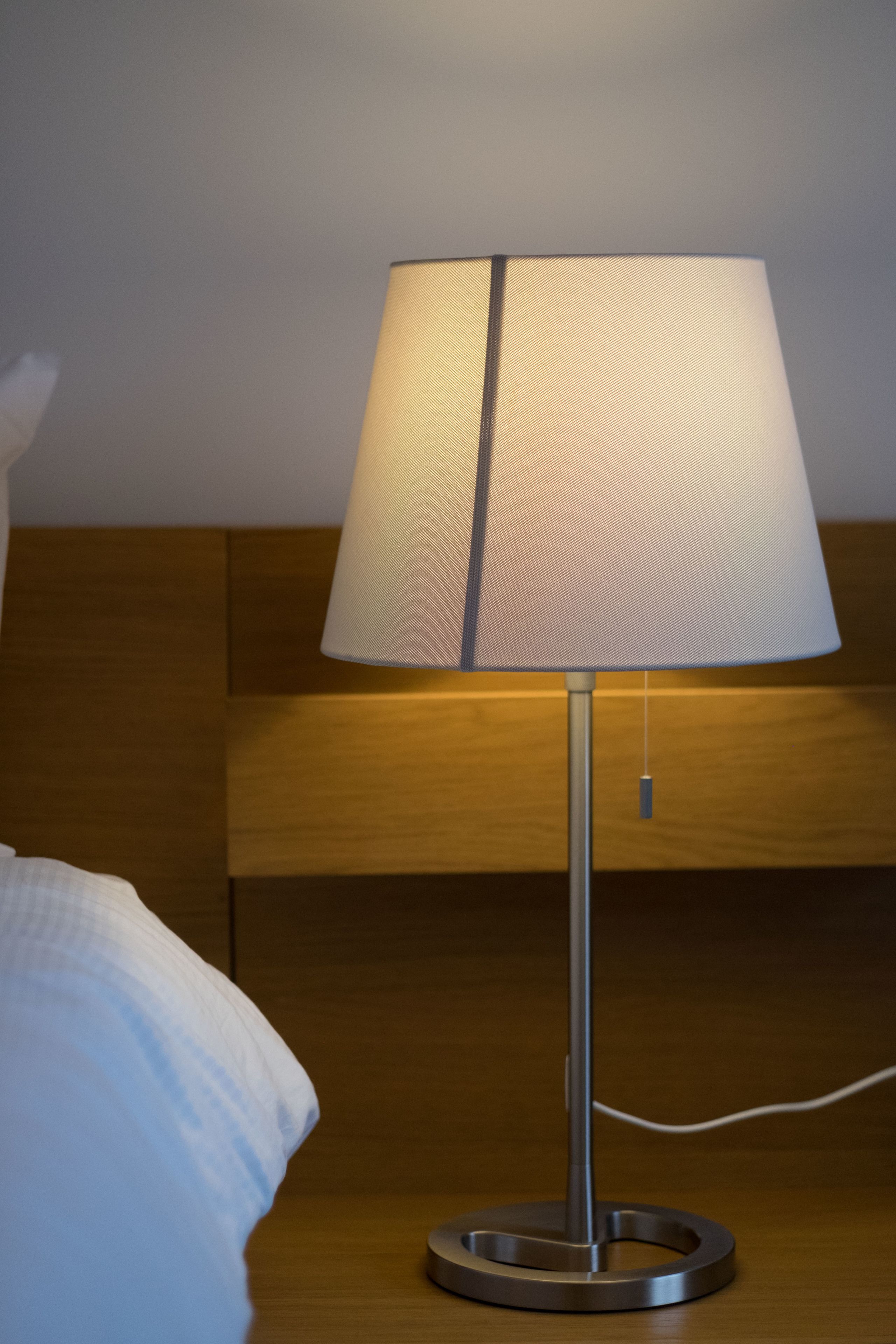 A modern table lamp with a metallic base and a fabric shade, placed on a wooden nightstand next to a bed with white bedding.