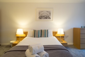 A hotel room with a bed, two bedside lamps, a wooden nightstand, a wooden dresser, and a framed picture on the wall.