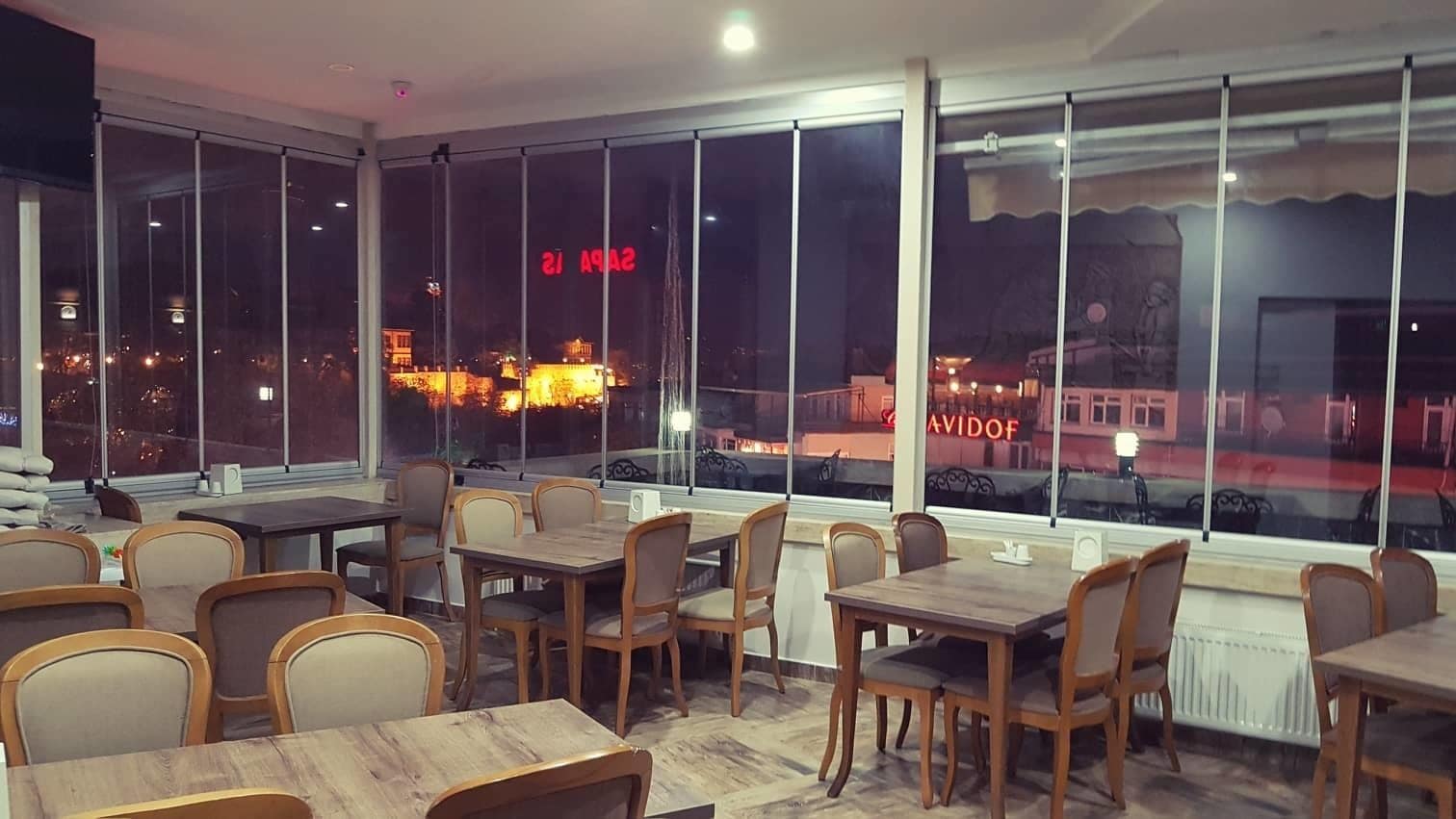 Een restaurant met meerdere tafels en stoelen, grote ramen met uitzicht op de stad 's nachts.
