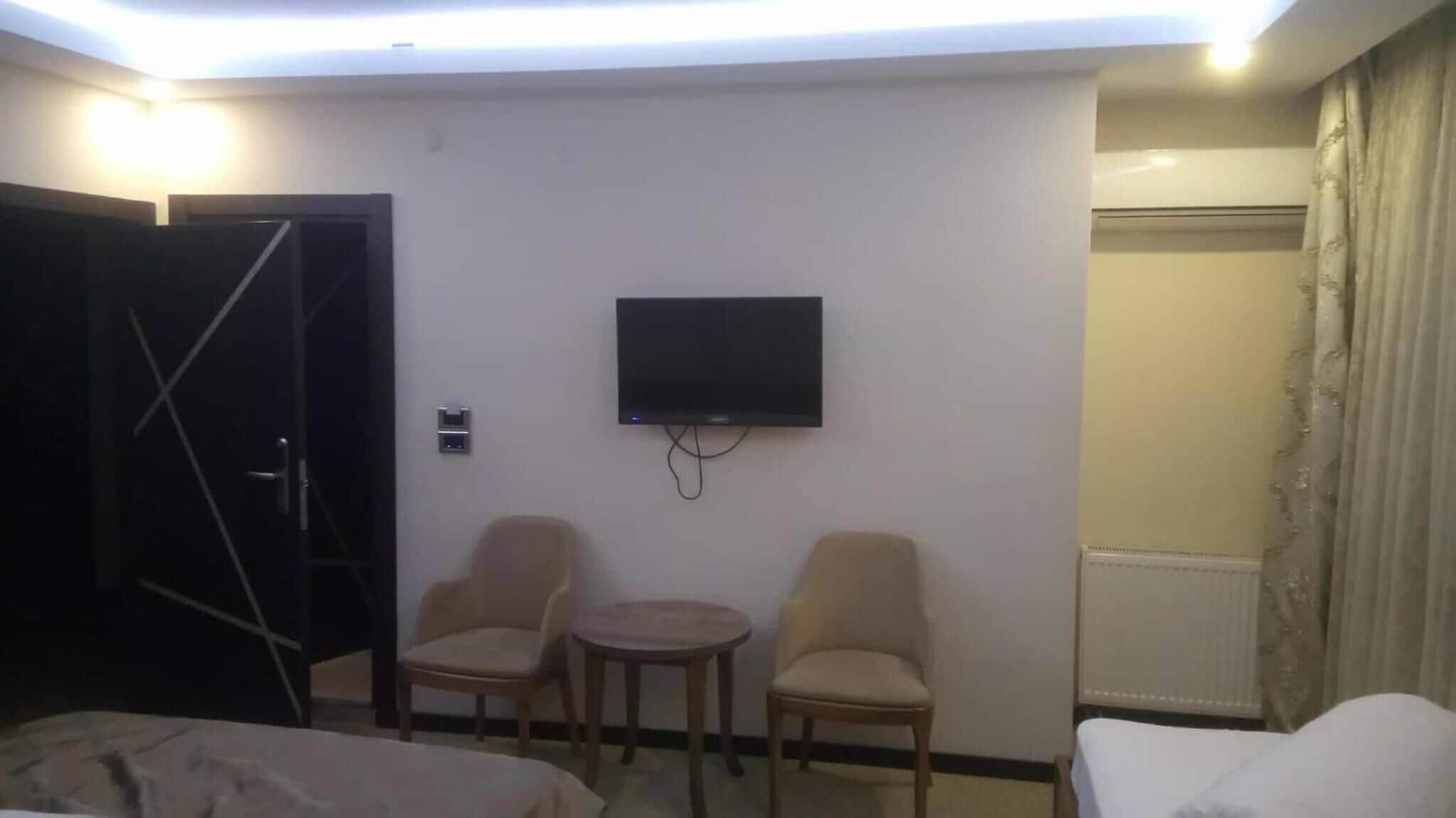 A hotel room with a flat-screen TV mounted on the wall, two chairs, a small table, a bed, and a door.