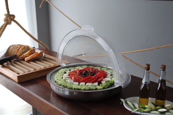 Una mesa de madera con un expositor de alimentos cubierto, pepinos en rodajas, tomates, queso, una barra de pan y dos botellas de aceite de oliva.
