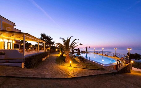 A pool area with a covered seating area and a view of the sea at dusk.
