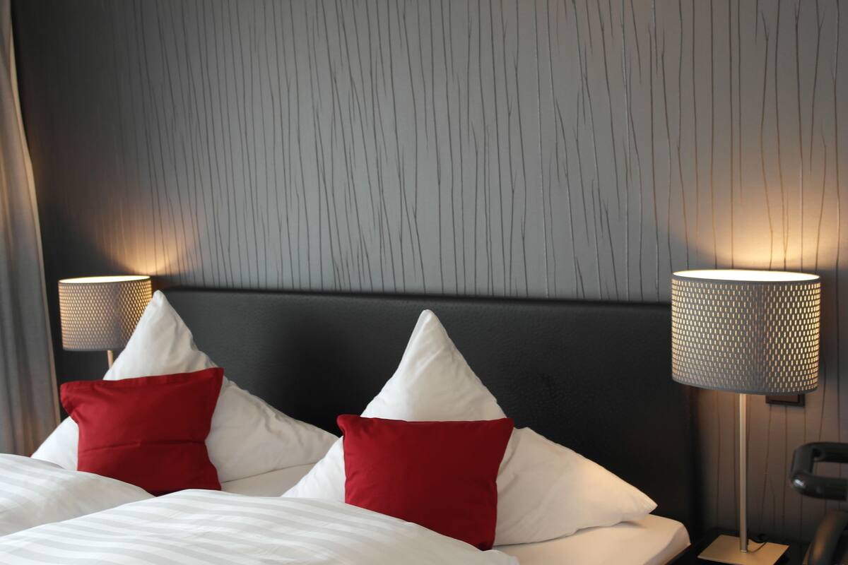 A hotel room with a bed, two red and white pillows, and two wall-mounted lamps.
