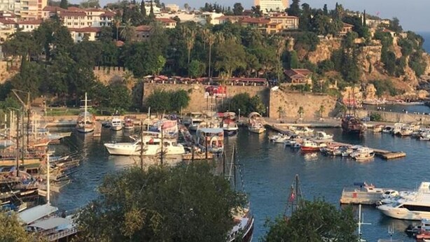 A coastal town with a marina filled with boats, a lighthouse, and buildings on a hillside. - Kont Pansiyon (Antalya)