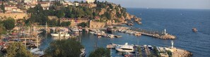 A coastal town with a marina filled with boats, a lighthouse, and buildings on a hillside. - Kont Pansiyon (Antalya)