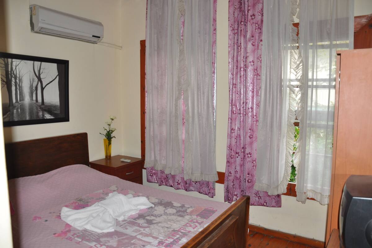 Une chambre à coucher comprenant un lit, une table de chevet, une lampe, une fenêtre avec des rideaux et un climatiseur.