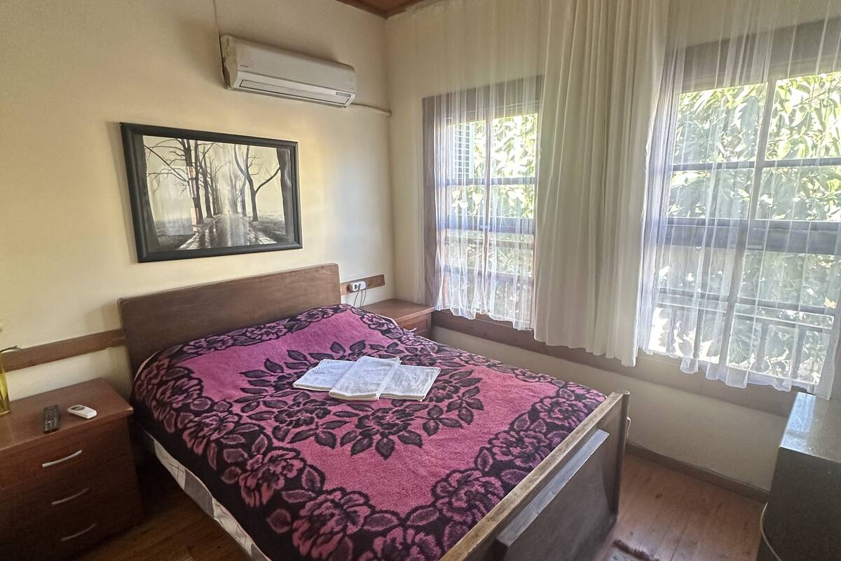 Une chambre avec un plafond en bois, un lit recouvert d’une couvre-lit à motifs floraux, une table de chevet, un climatiseur fixé au mur et une fenêtre ornée de rideaux légers.